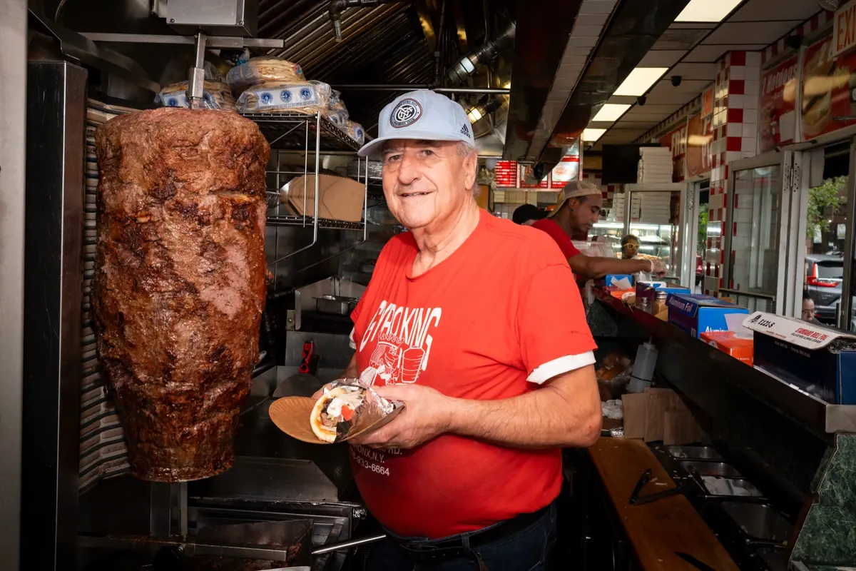 The owner at the gyro spit, holding a fresh gyro, in a red Gyro King shirt
